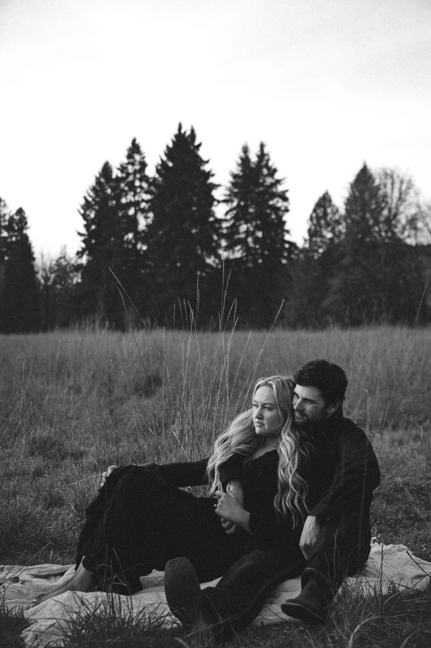 A couple sits together in a grassy field with evergreen trees, captured in moody black and white photography.