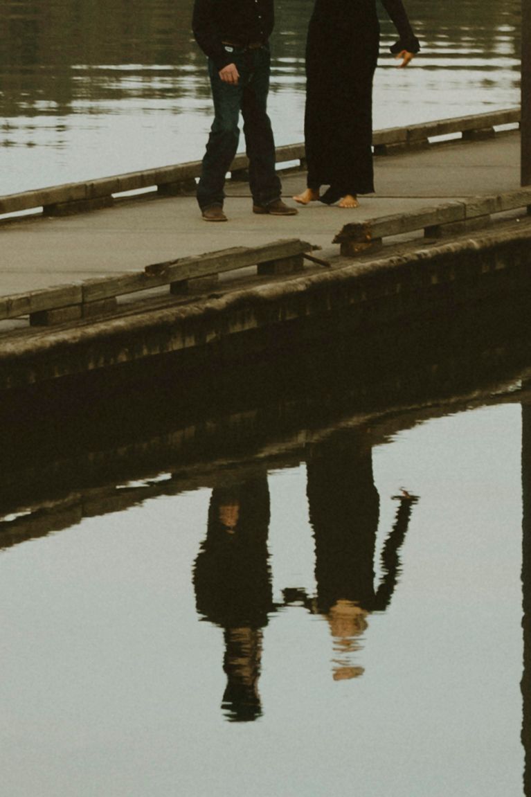 Two people standing on a wooden dock reflected in calm water below, captured in moody tones.