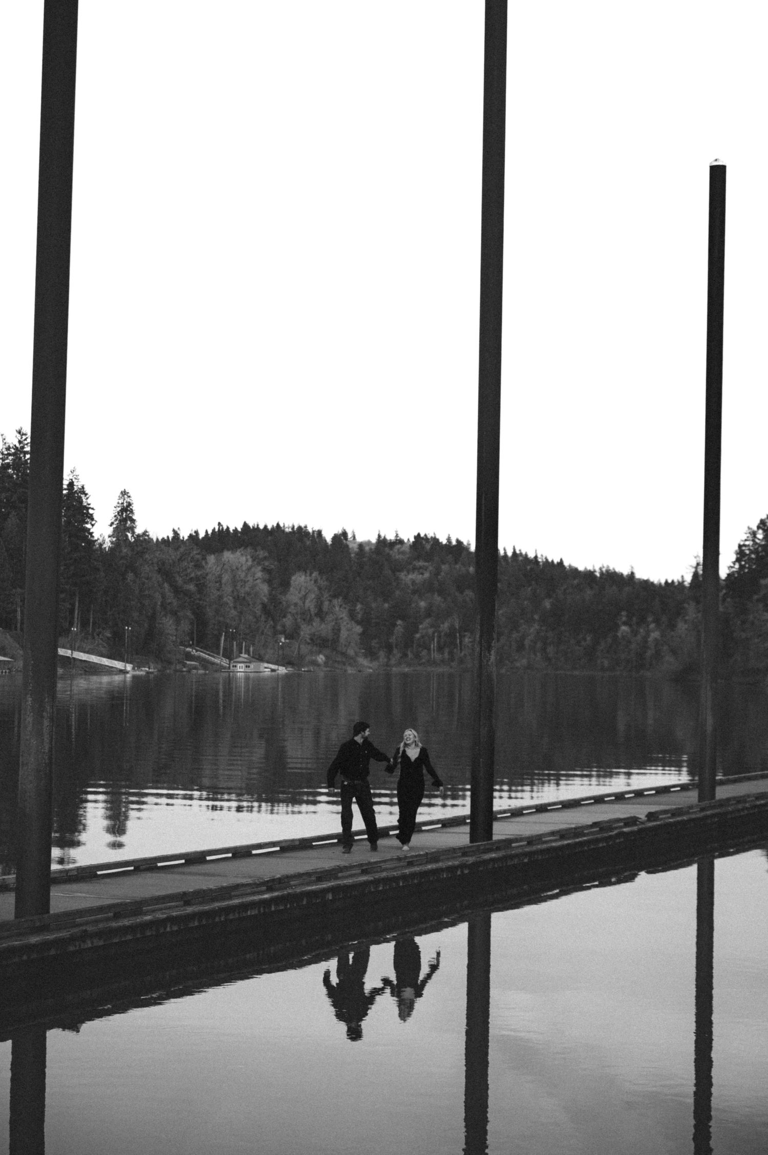Black and white photo of a couple holding hands on a reflective dock by a serene lake surrounded by tall trees.
