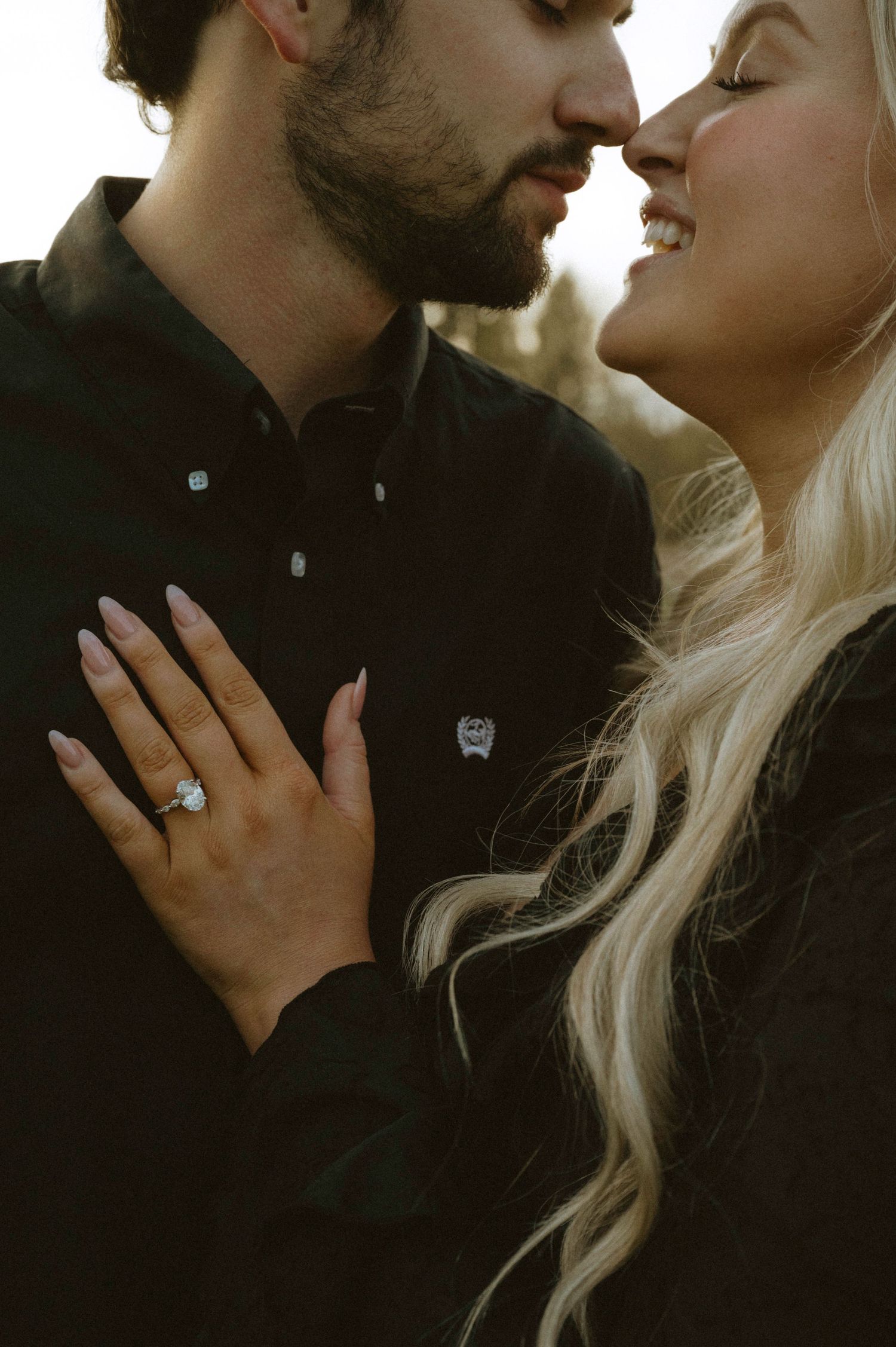 Couple in black outfits sharing a romantic moment, woman displaying a diamond engagement ring on her hand.