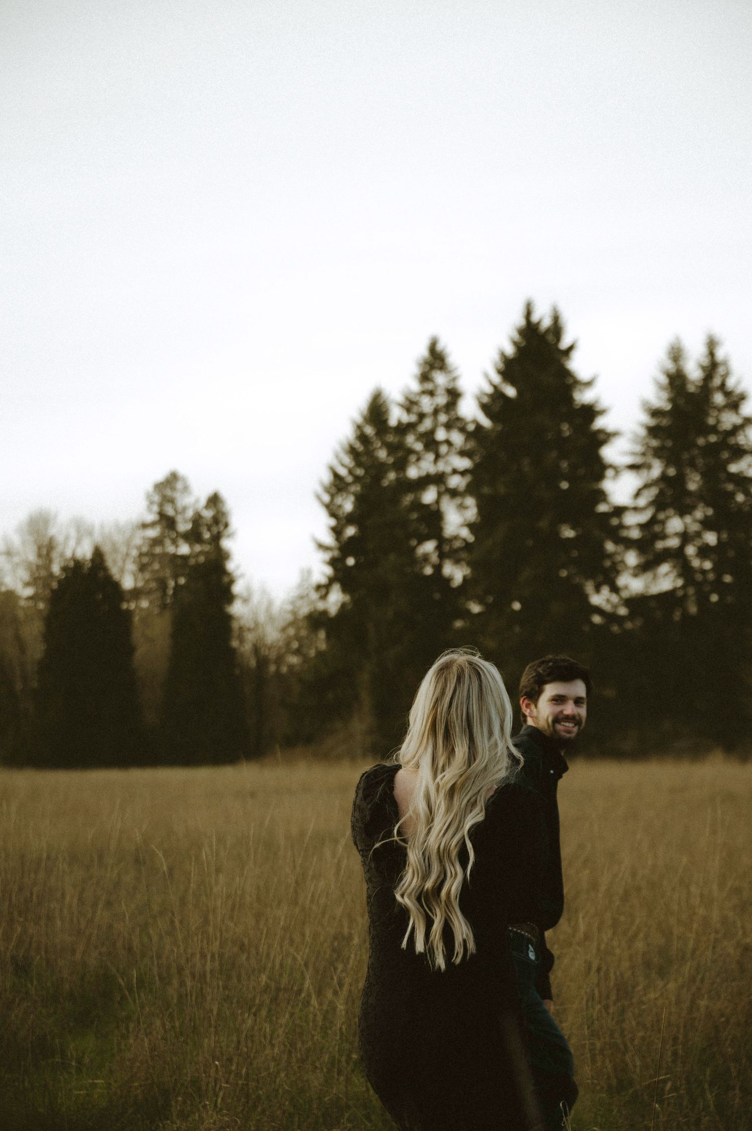 Couple embracing in golden field with evergreen trees, woman with long blonde hair in black outfit.