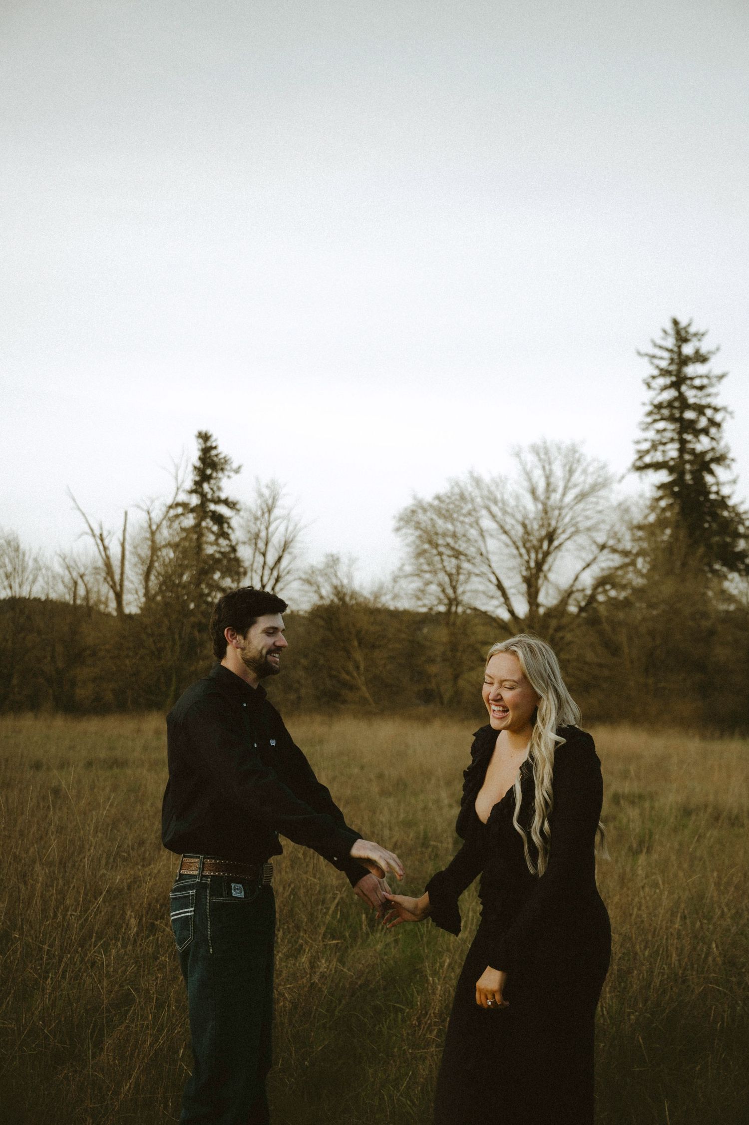 A couple in black outfits holds hands and laughs together in a golden grass field with bare trees at dusk.