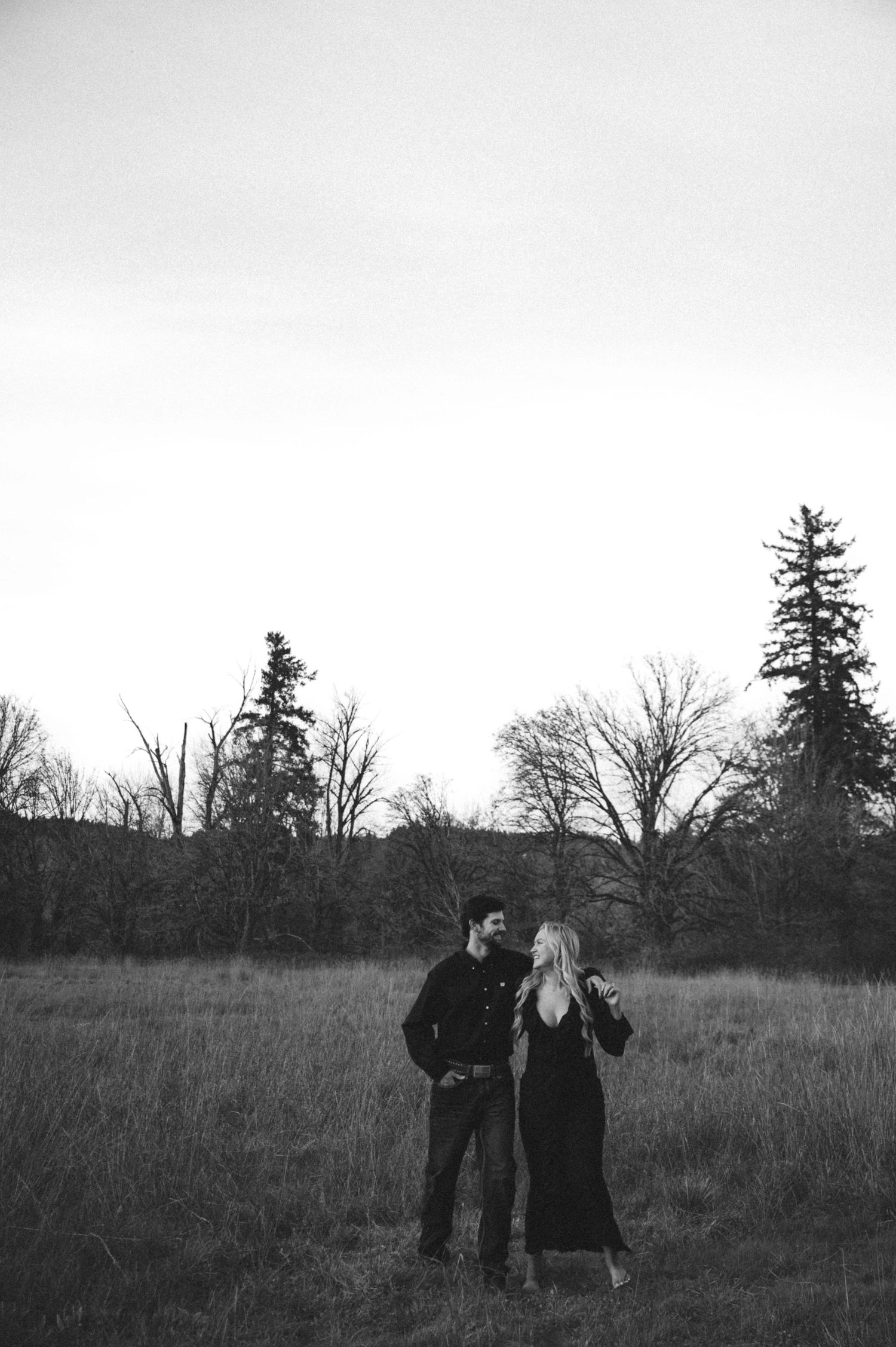 Black and white photo of couple standing in field with bare trees, sharing a romantic moment.