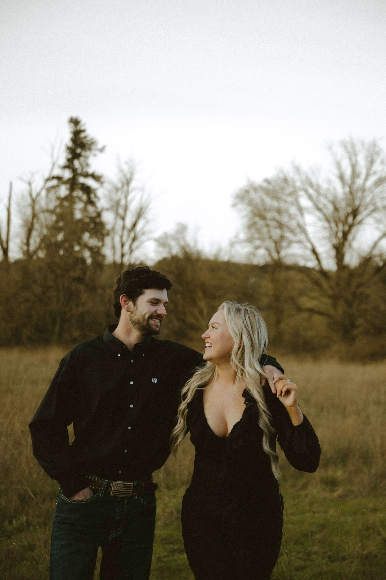 A smiling couple in matching black outfits laughing together in a rustic field with bare trees in the background.