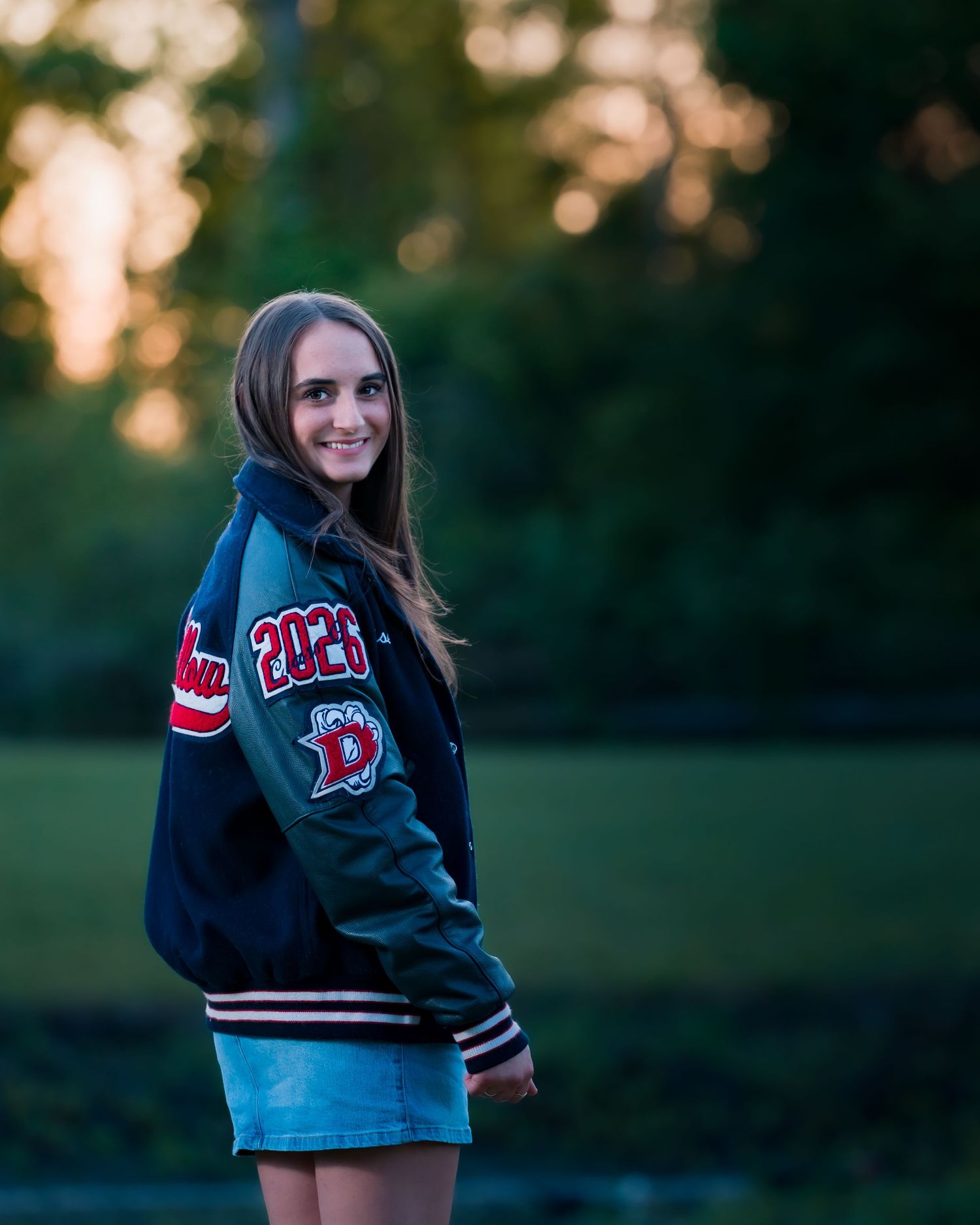 Teen girl smiling in a varsity letterman jacket with 2026 patch, standing outdoors at golden hour.