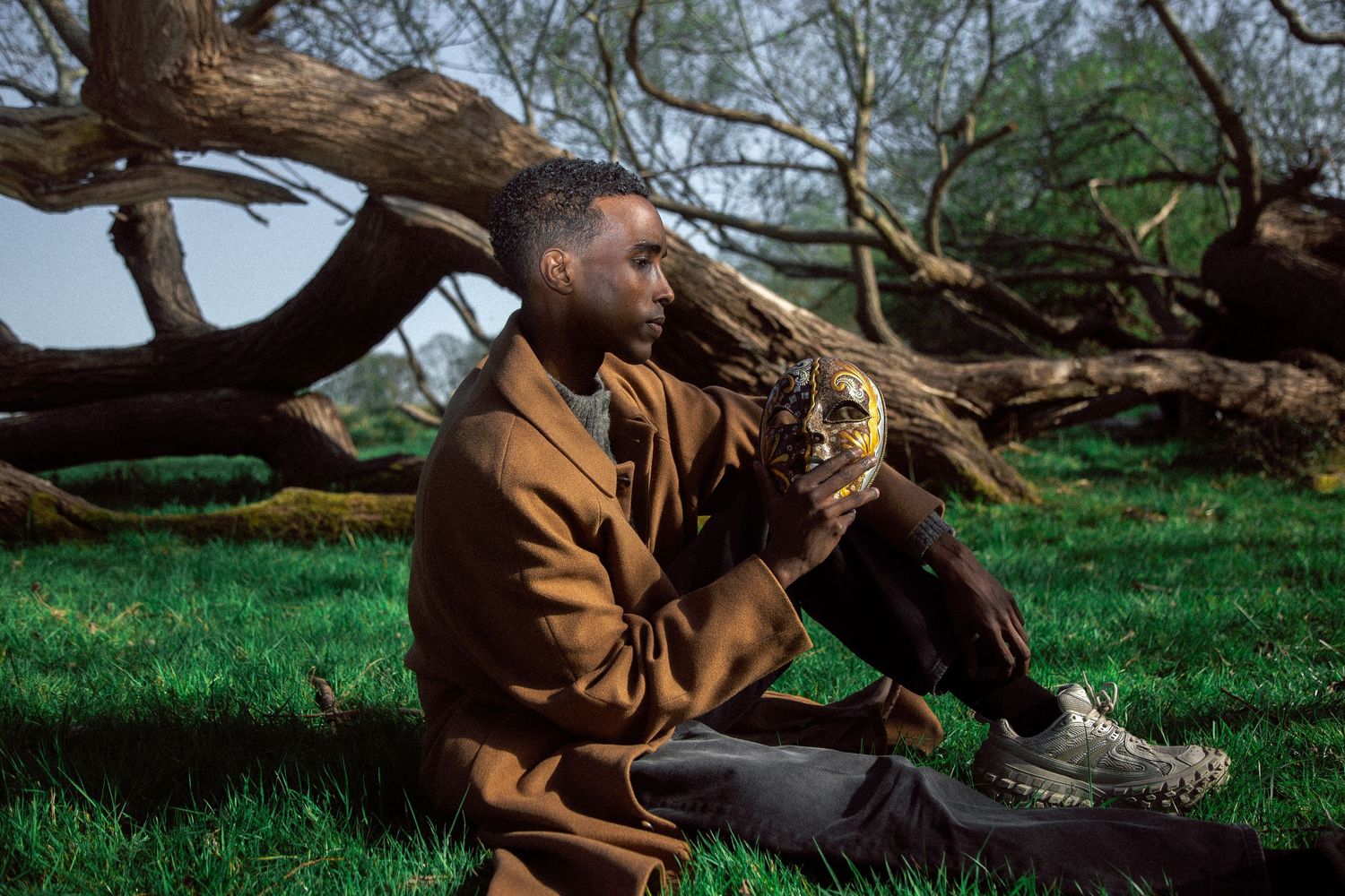Man in tan blazer holding ornate decorative mask while sitting outdoors near fallen trees on green grass.