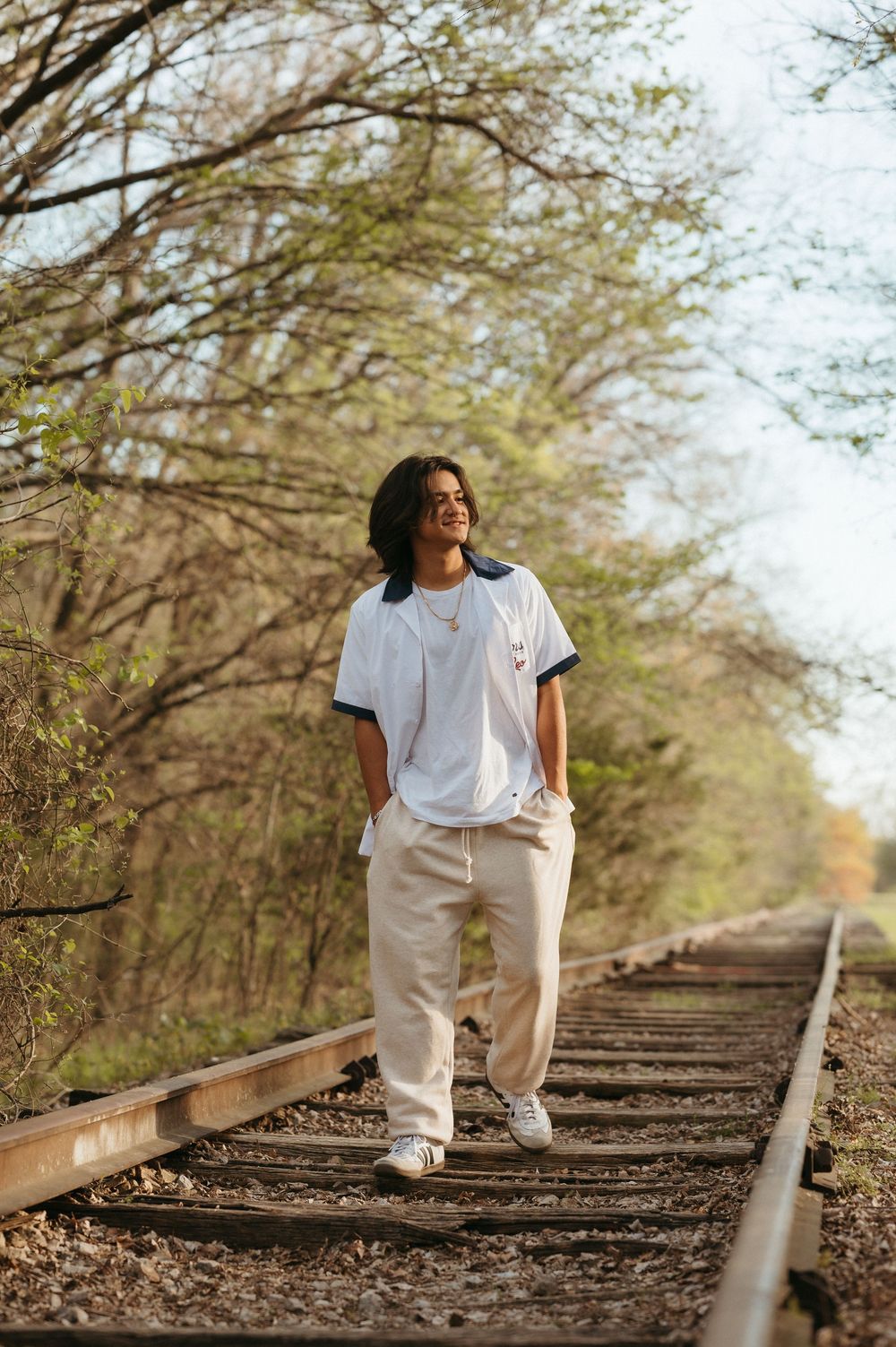 Young man with curly hair smiling on railroad tracks, wearing white tee and beige pants, surrounded by spring trees.
