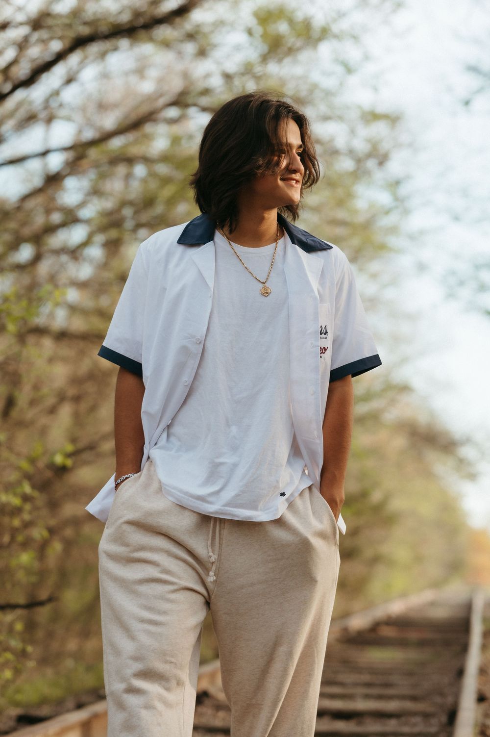 Young man in white shirt and beige pants standing on railroad tracks surrounded by trees.