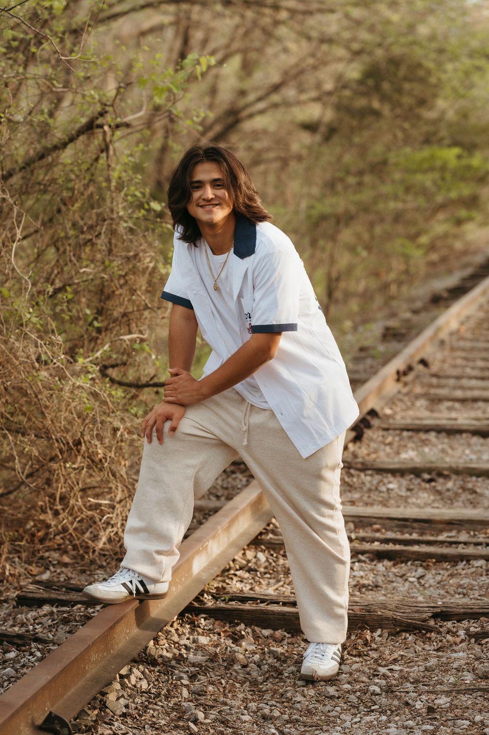 Young person in white polo and cream pants posing on railway tracks surrounded by trees.