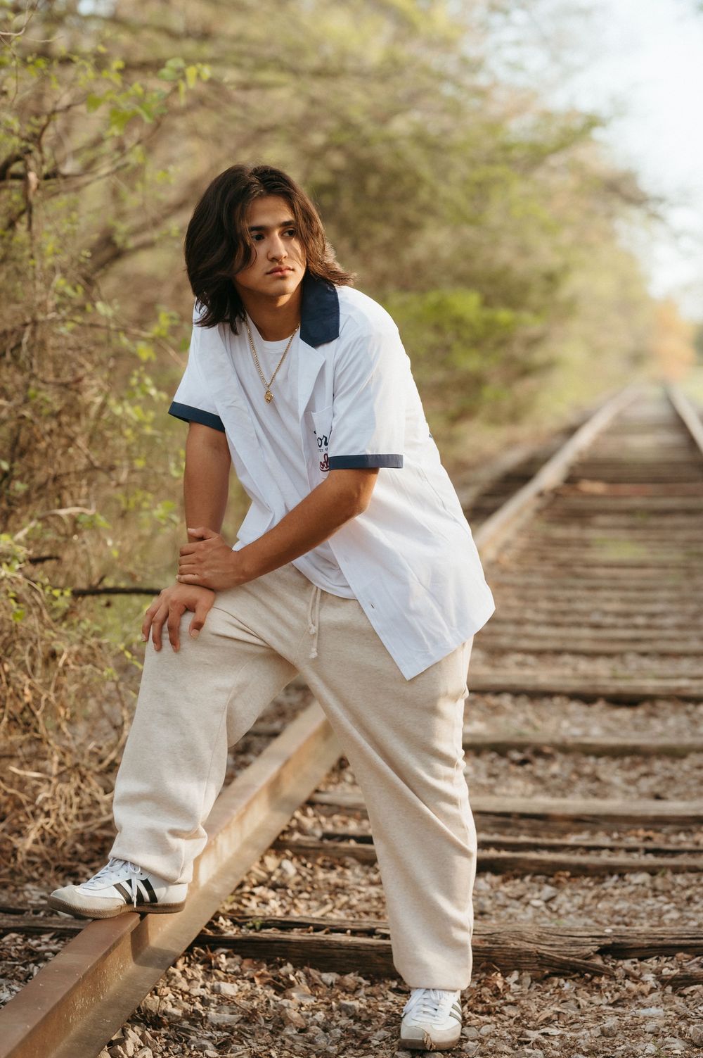 Young man with long hair posing on railroad tracks, wearing white shirt and cream pants with Adidas sneakers.