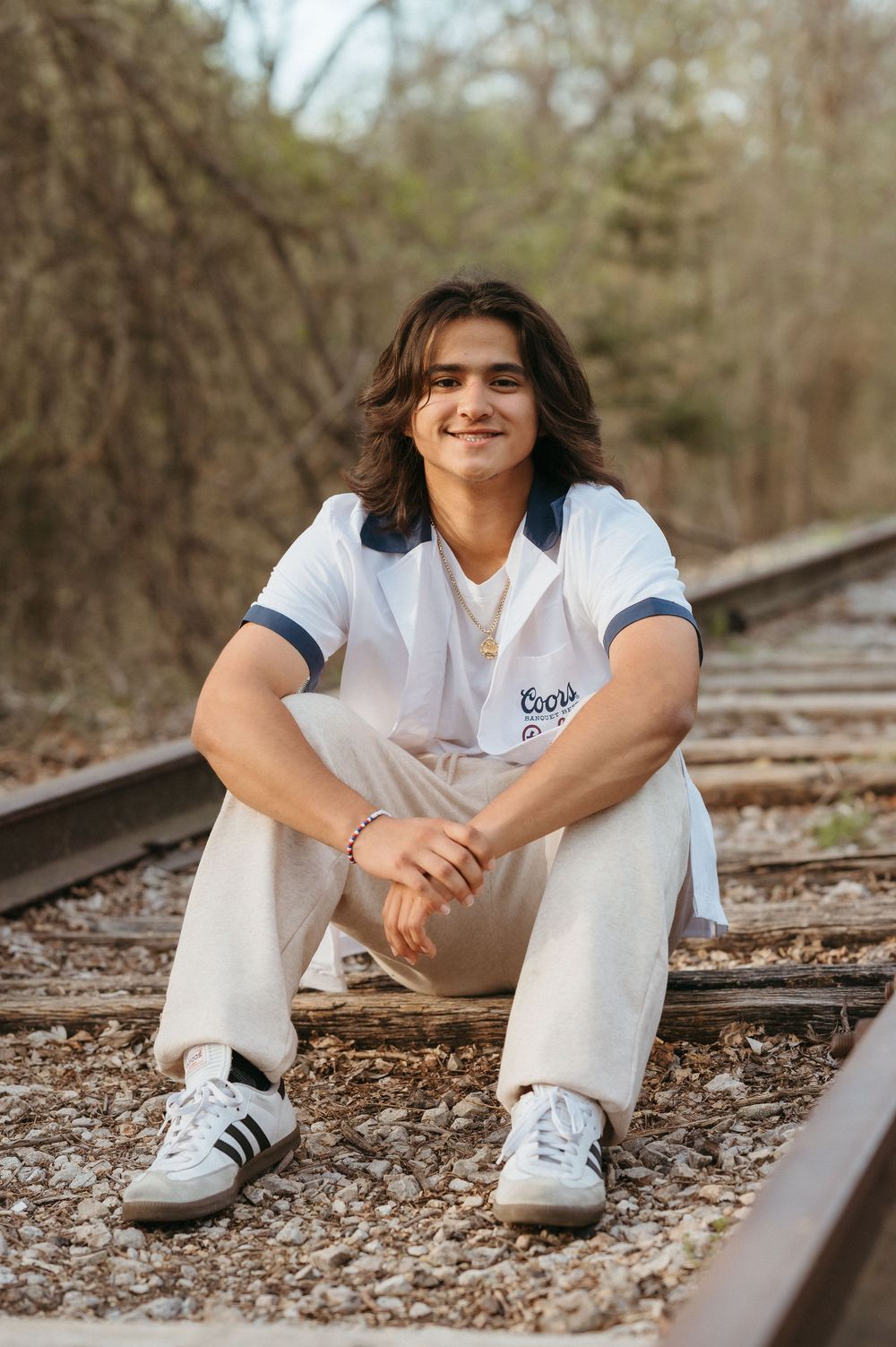 Young man with long hair sitting on railroad tracks wearing white outfit and Adidas sneakers, smiling outdoors.
