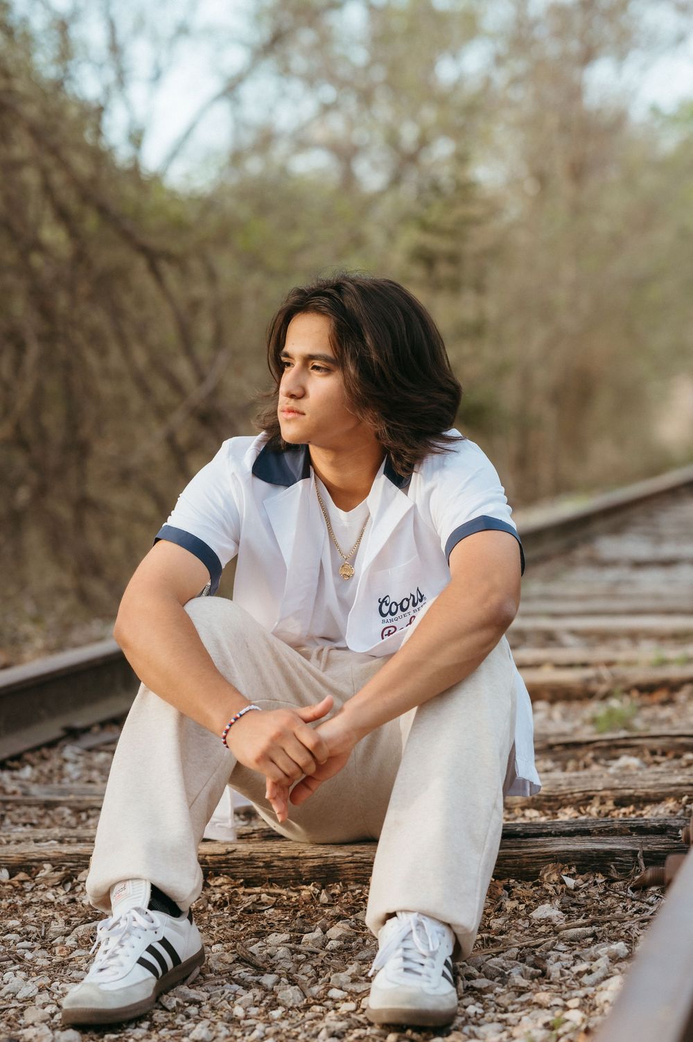 Young man with long dark hair sits on railroad tracks wearing white Coors shirt, cream pants, and Adidas sneakers.