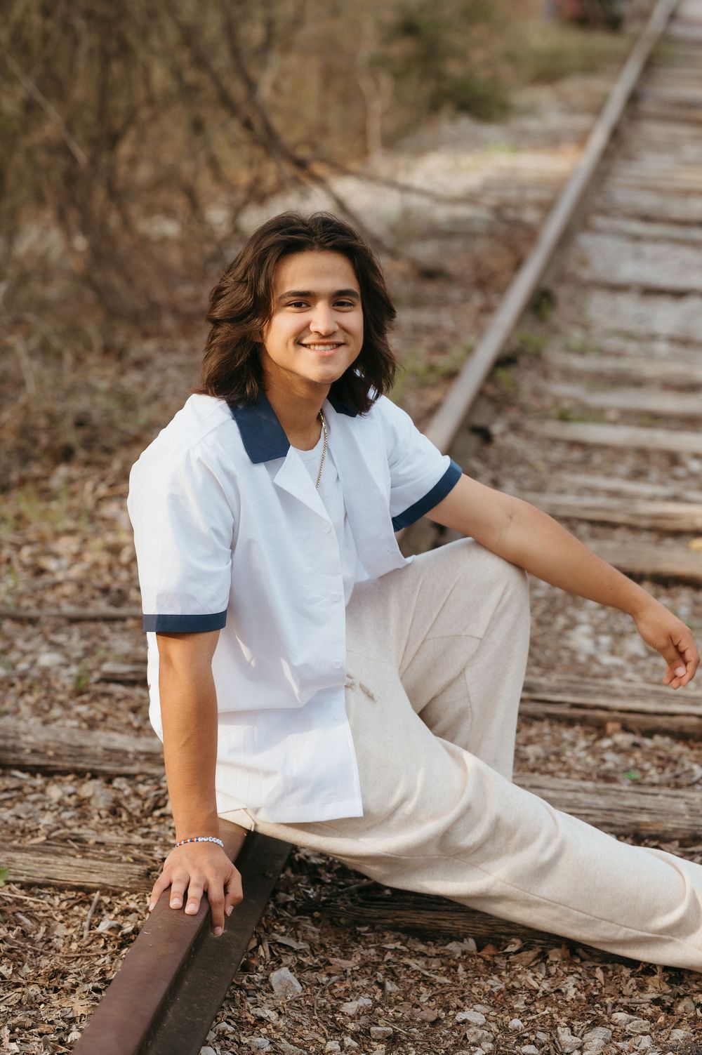 Smiling young person with long hair sits on railroad tracks wearing white polo shirt and beige pants outdoors.