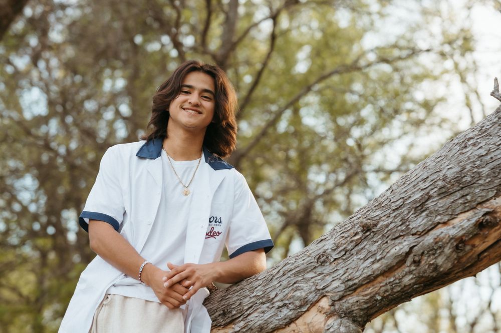 Young person smiling outdoors, leaning against a tree trunk, wearing a white top with blue trim.