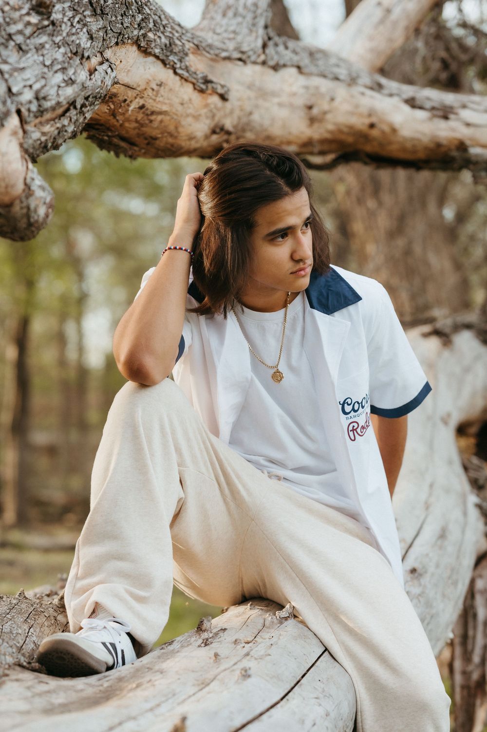 Young man with long hair sitting on driftwood in forest, wearing white tee and cream pants with gold chain.