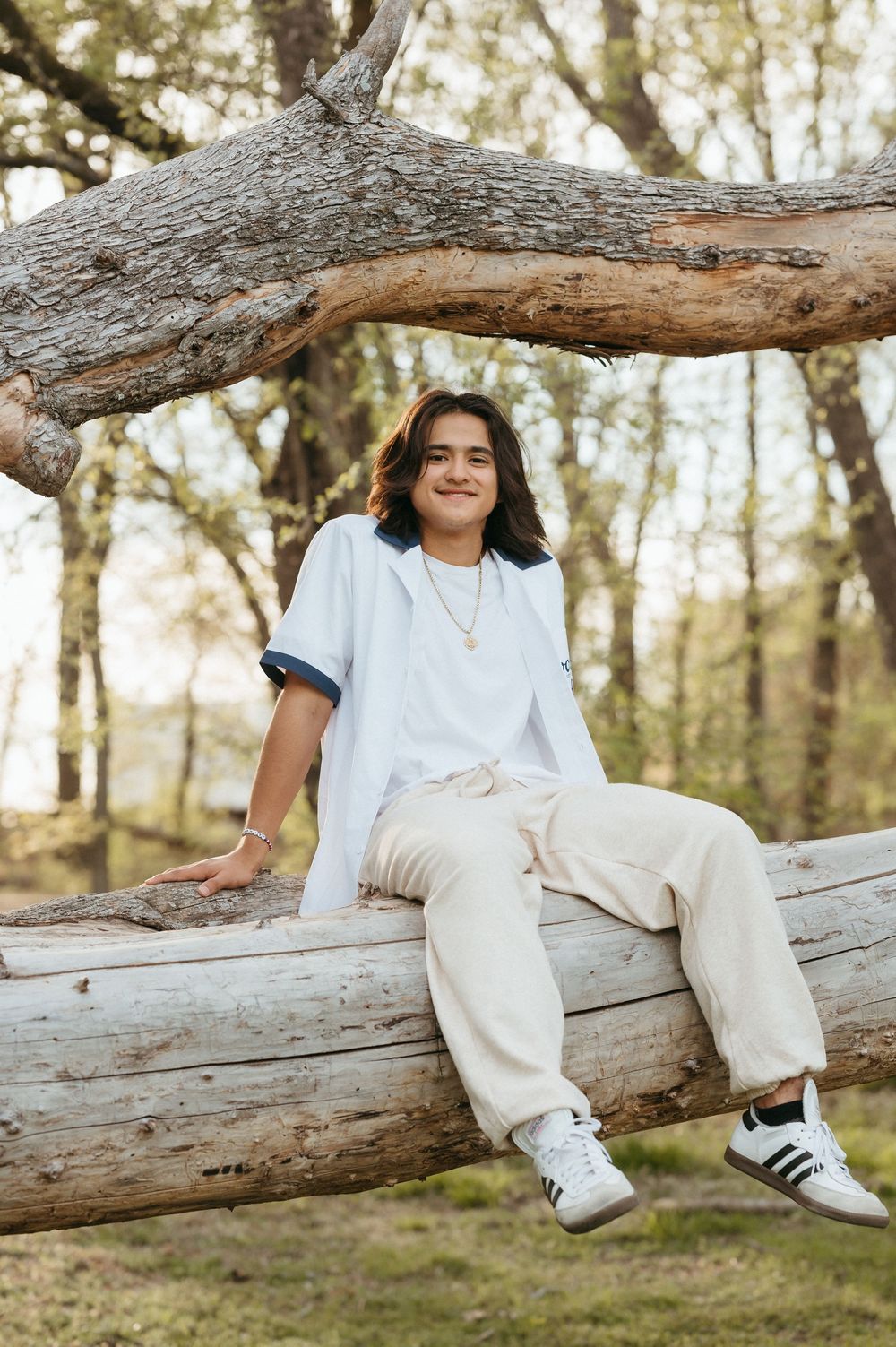 Teen boy with long hair sits smiling on fallen log in wooded park wearing white shirt and cream pants.