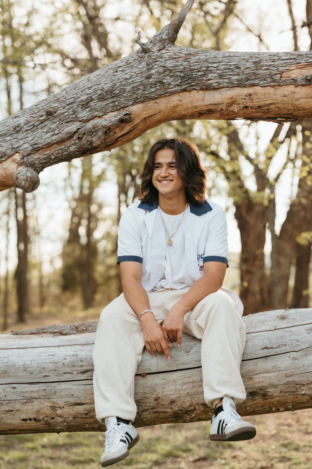 Smiling young man with long hair sitting on a fallen log in a forest, wearing white outfit and Adidas sneakers.