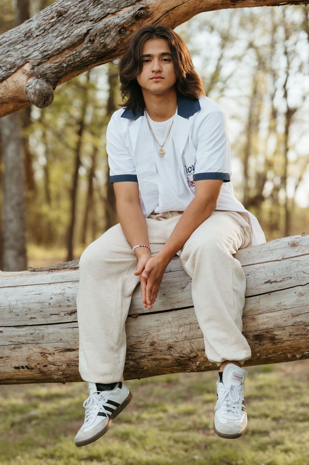 Young man with long hair sits on a log in a forest, wearing a white polo, cream pants, and Adidas sneakers.
