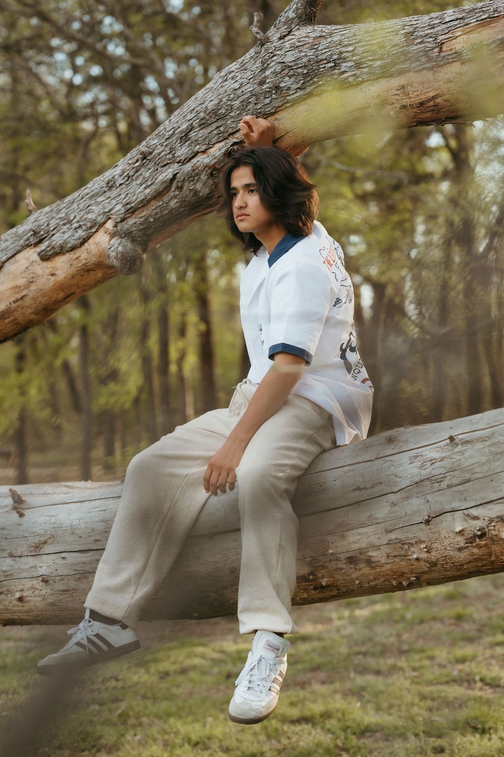 Young person sitting on a fallen log in a forest, wearing a white shirt and beige pants with sneakers.