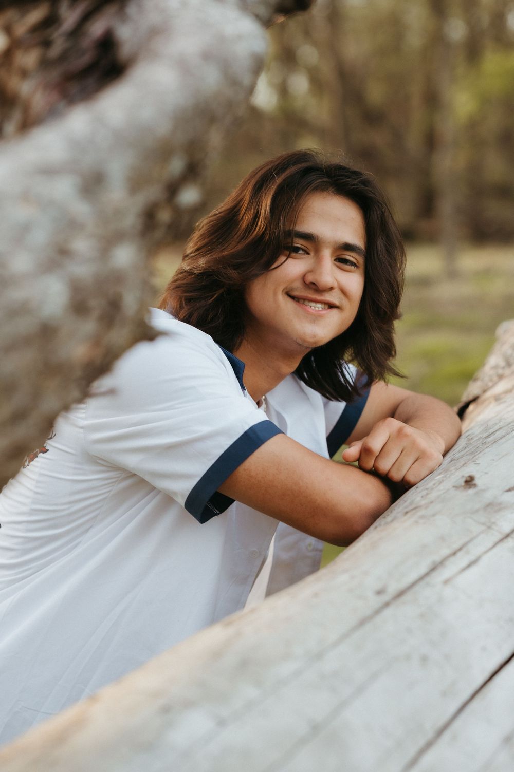 Smiling young man with long brown hair leaning on a wooden log in a park, wearing a white shirt.
