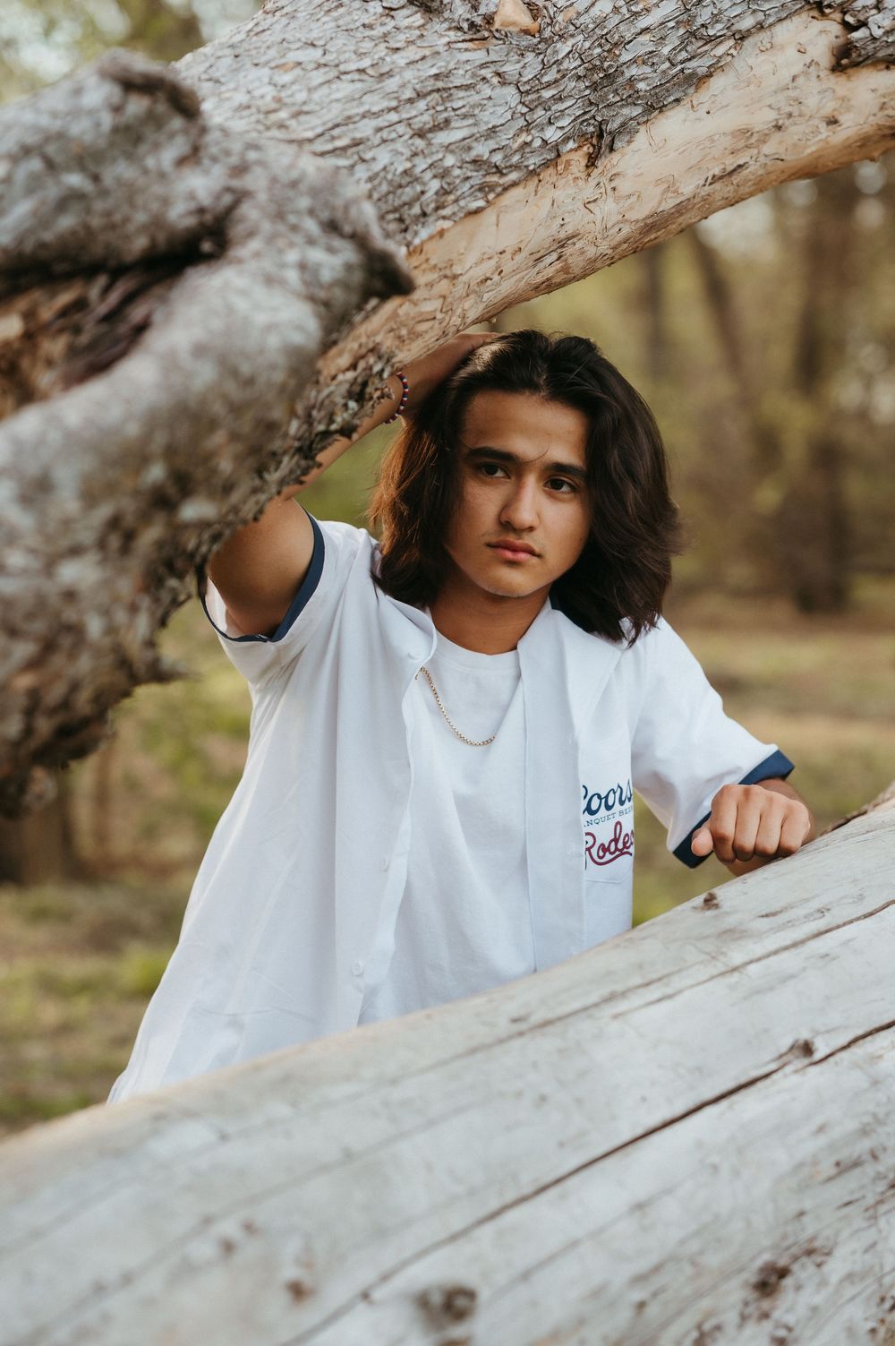 Young man with long hair in white graphic tee poses between fallen logs in a forest setting.