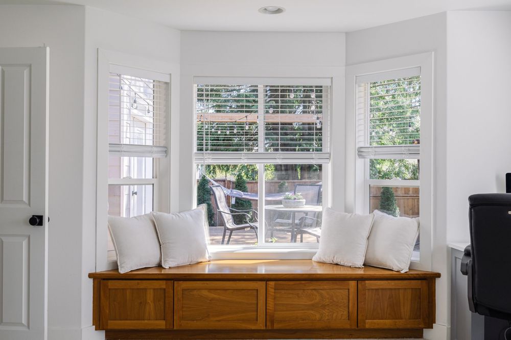 Bright bay window seat with white cushions, wood storage bench, and plantation shutters.