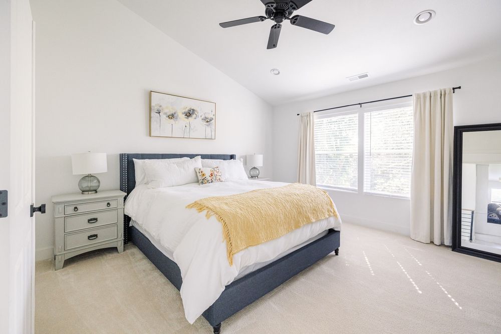 Elegant master bedroom with navy upholstered bed, yellow throw, ceiling fan, and large bright window.