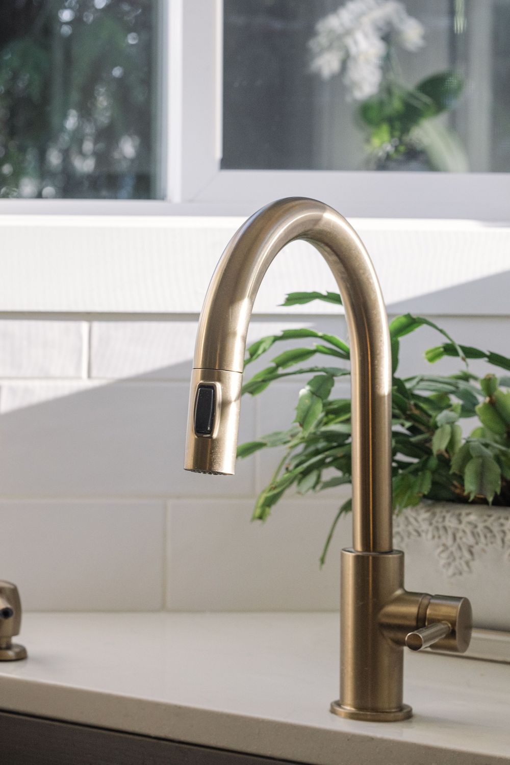 Brushed gold pull-down kitchen faucet on white countertop with subway tile backsplash and green plants.