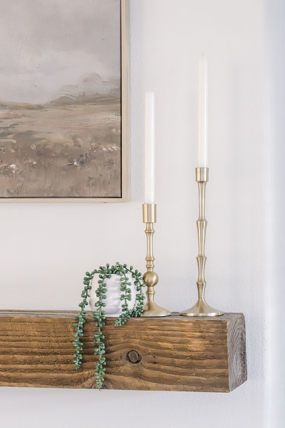 Rustic wooden shelf with gold candlesticks, trailing succulent, and neutral landscape art on white wall.
