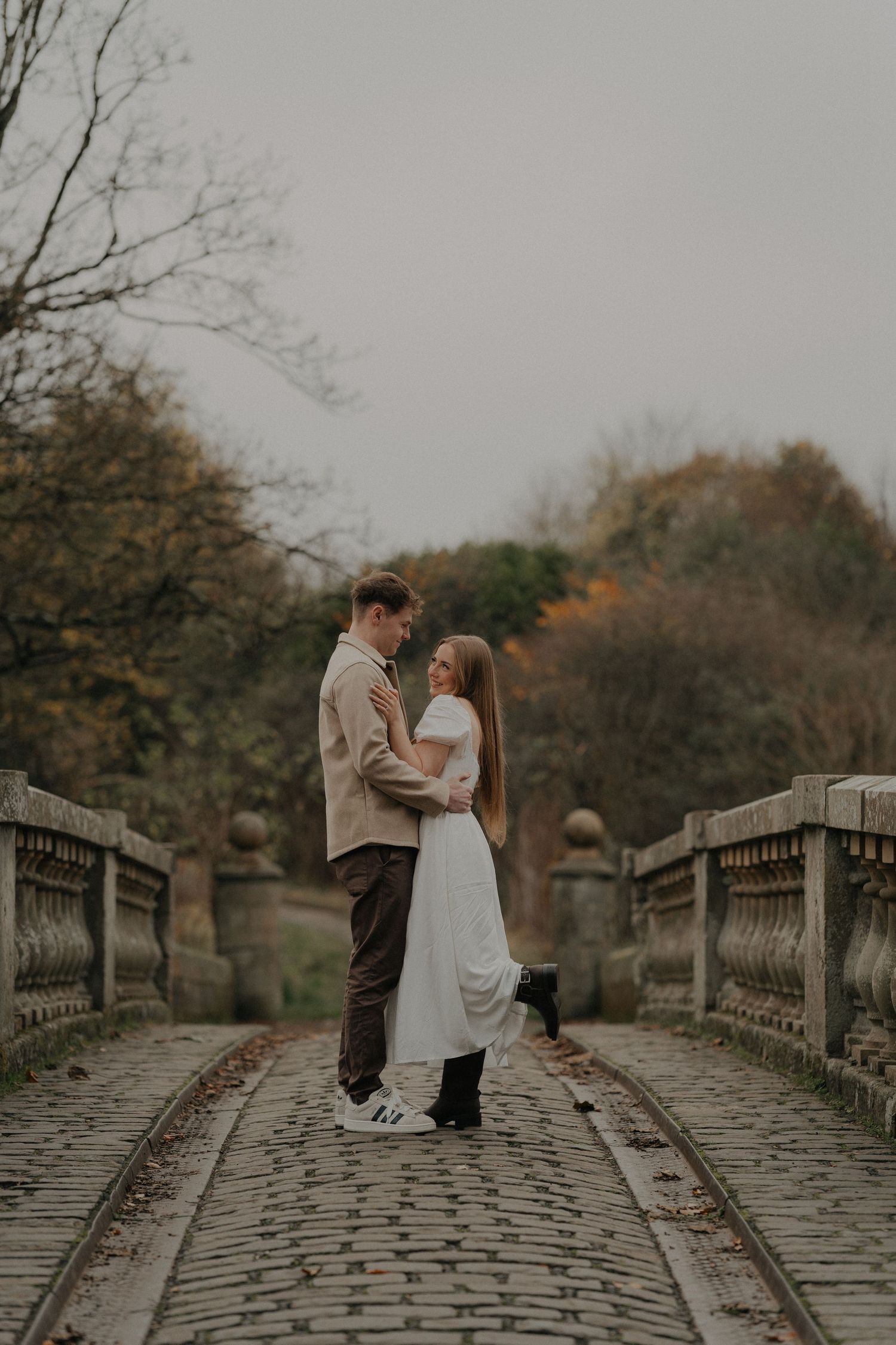 A couple embraces tenderly under a wooden gazebo, surrounded by autumn foliage, as the man holds the woman in a white dress.