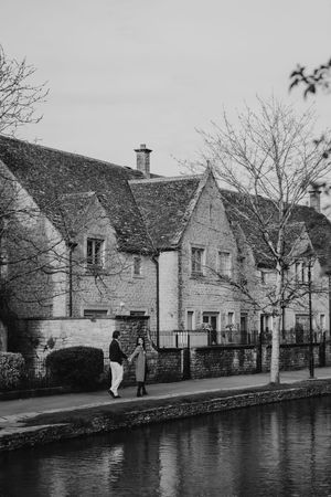 Couple walking along a canal beside historic stone cottages in a charming English village in bourton on the water, black and white photo.