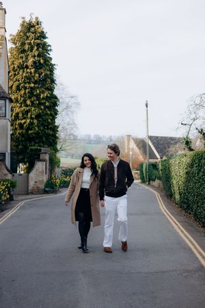 Couple walking hand in hand down at their Cotswolds engagement photoshoot, surrounded by stone buildings and lush greenery on a bright day.
