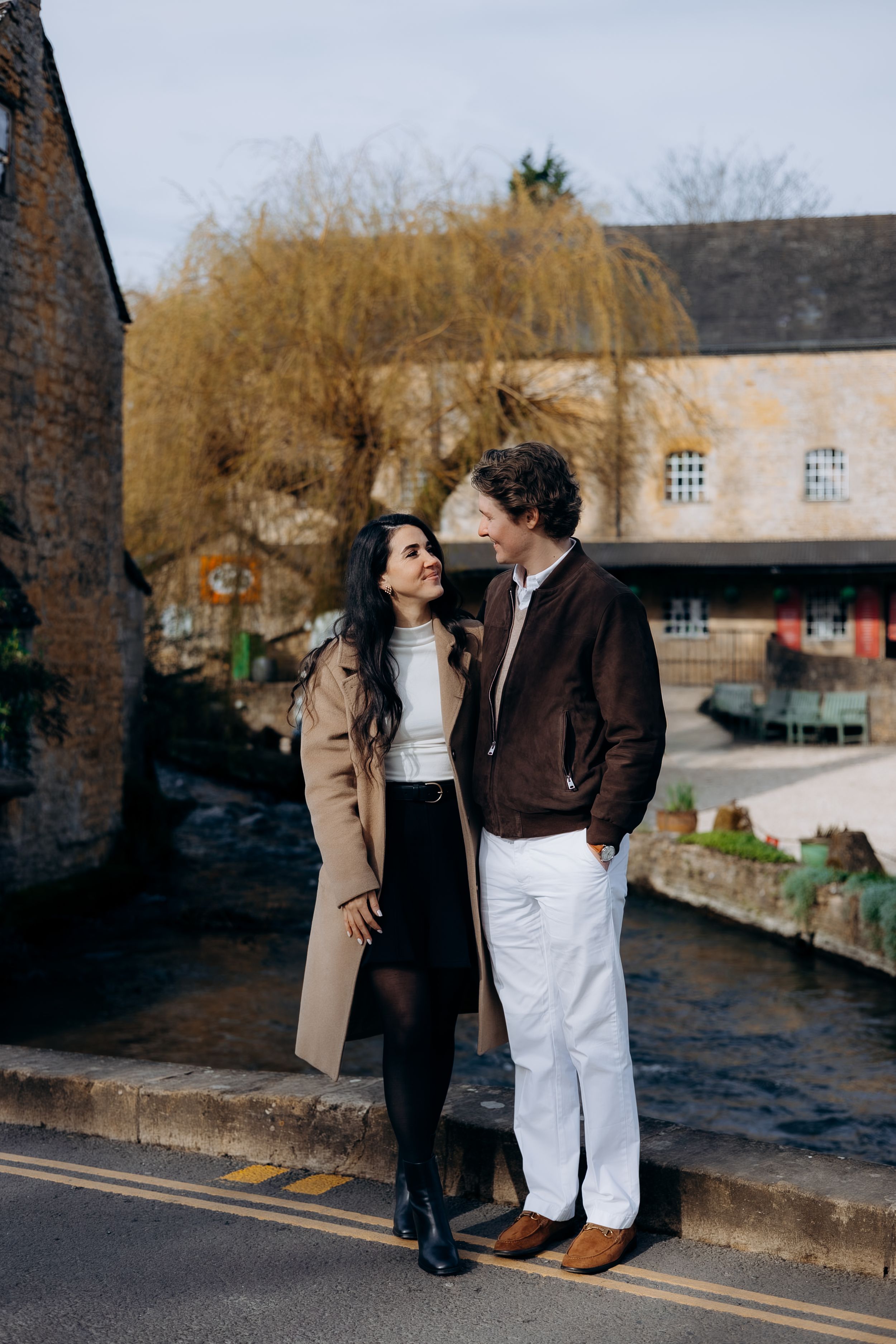 A couple stands together on a cobblestone street in a charming cotswolds village, smiling at each other near a stream.