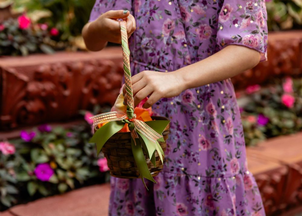 Girl in purple floral dress holding a decorative woven basket with green ribbon bow in a garden setting.