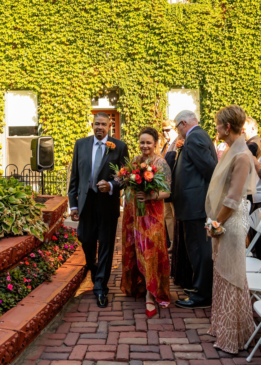 Bride in colorful gown walks down brick aisle with escort at outdoor wedding ceremony beside ivy-covered wall.