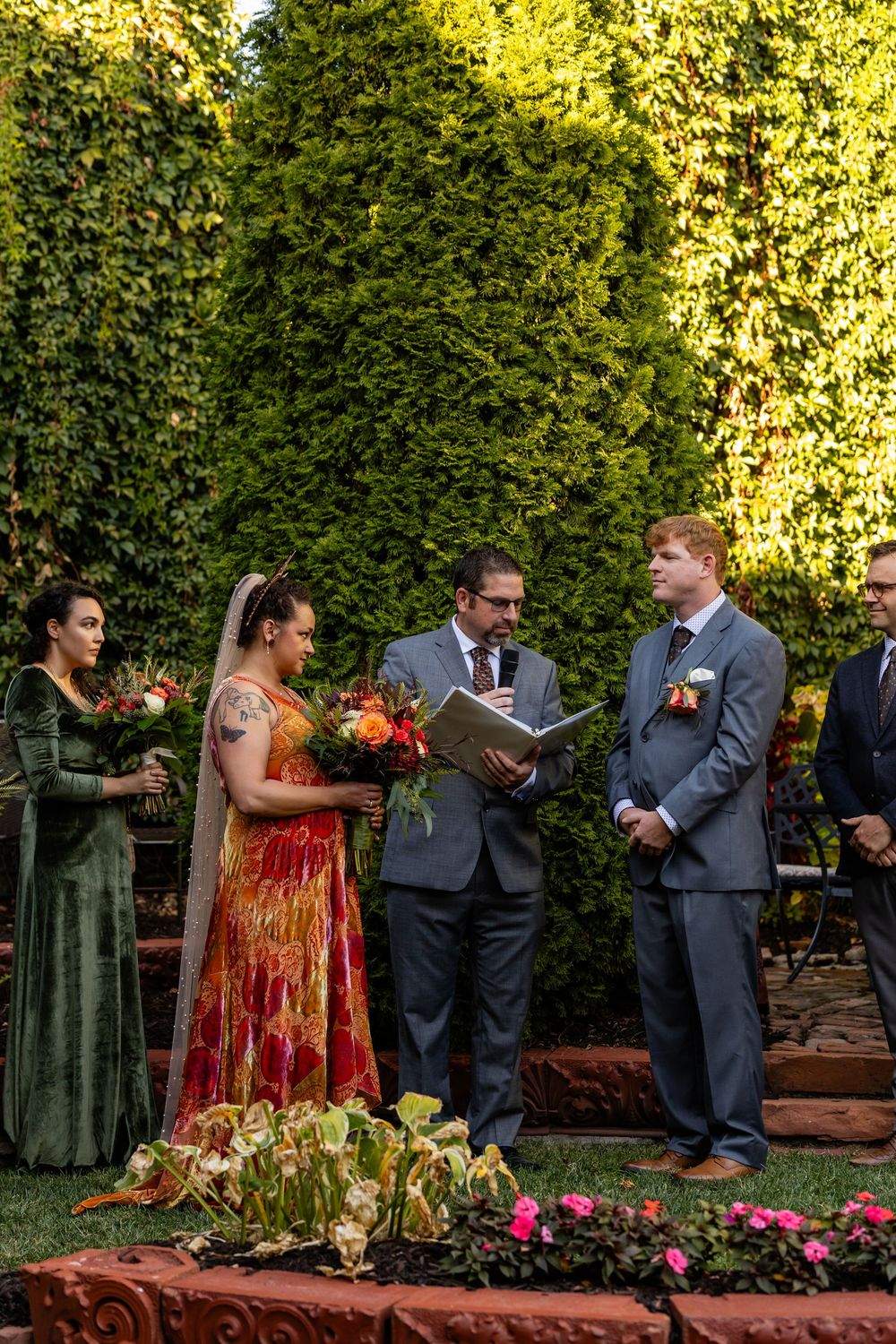 Bride in red dress and groom in gray suit exchange vows with officiant in lush garden ceremony.
