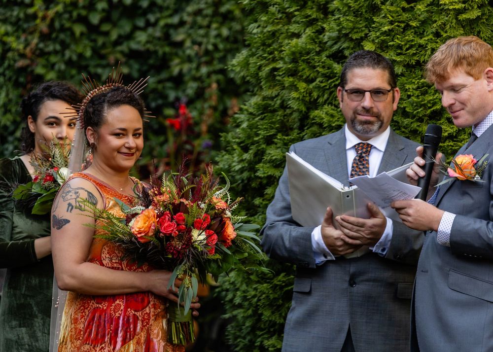 Outdoor wedding ceremony with bride holding colorful bouquet, officiant reading vows, and guests smiling against green ivy backdrop.