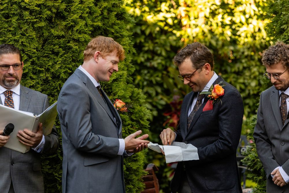 Two grooms exchange rings during an outdoor wedding ceremony surrounded by lush greenery and orange floral arrangements.