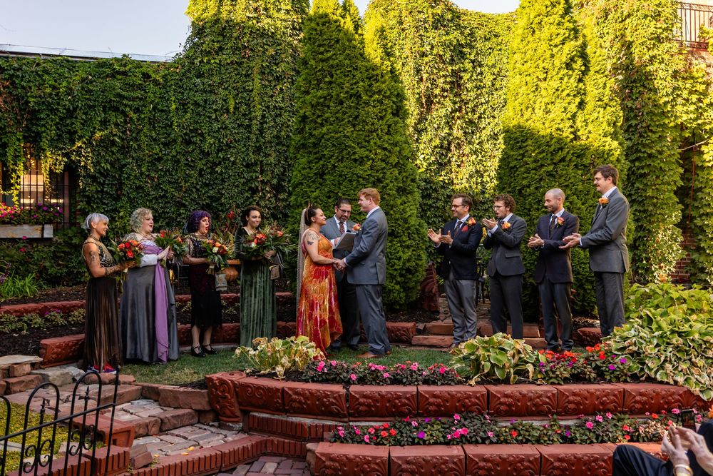 Wedding party gathered in lush green garden ceremony, bride in colorful gown exchanging vows with groom surrounded by attendants.