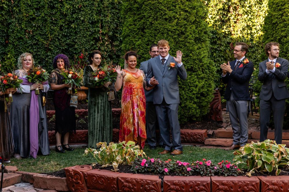 Wedding party celebrates outdoors, bride in colorful dress stands with groom and attendants in garden setting.