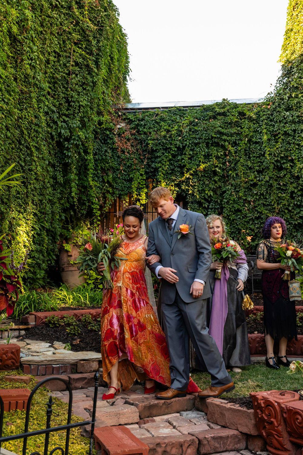 Newlywed couple walks together through garden venue after outdoor wedding ceremony with bridal party.