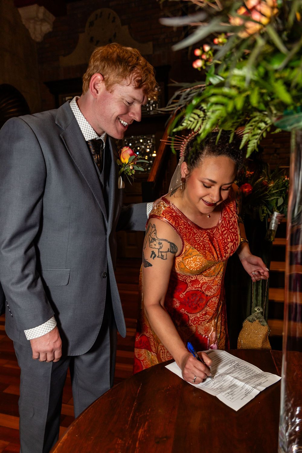 Bride in red dress signs wedding certificate while groom in gray suit smiles beside her at wooden table.