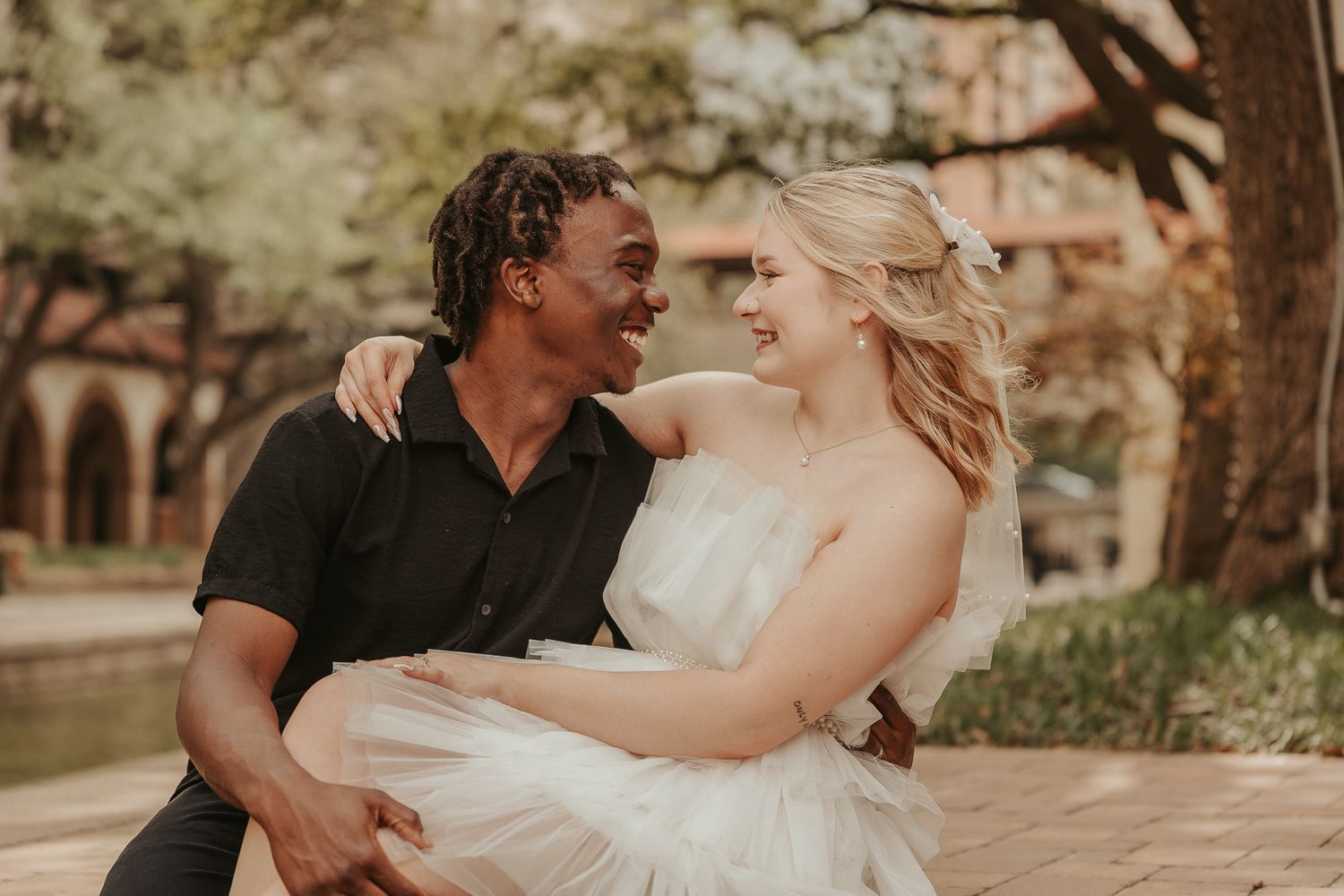 Joyful interracial couple sharing a happy moment outdoors, bride in white dress held by groom in black shirt.