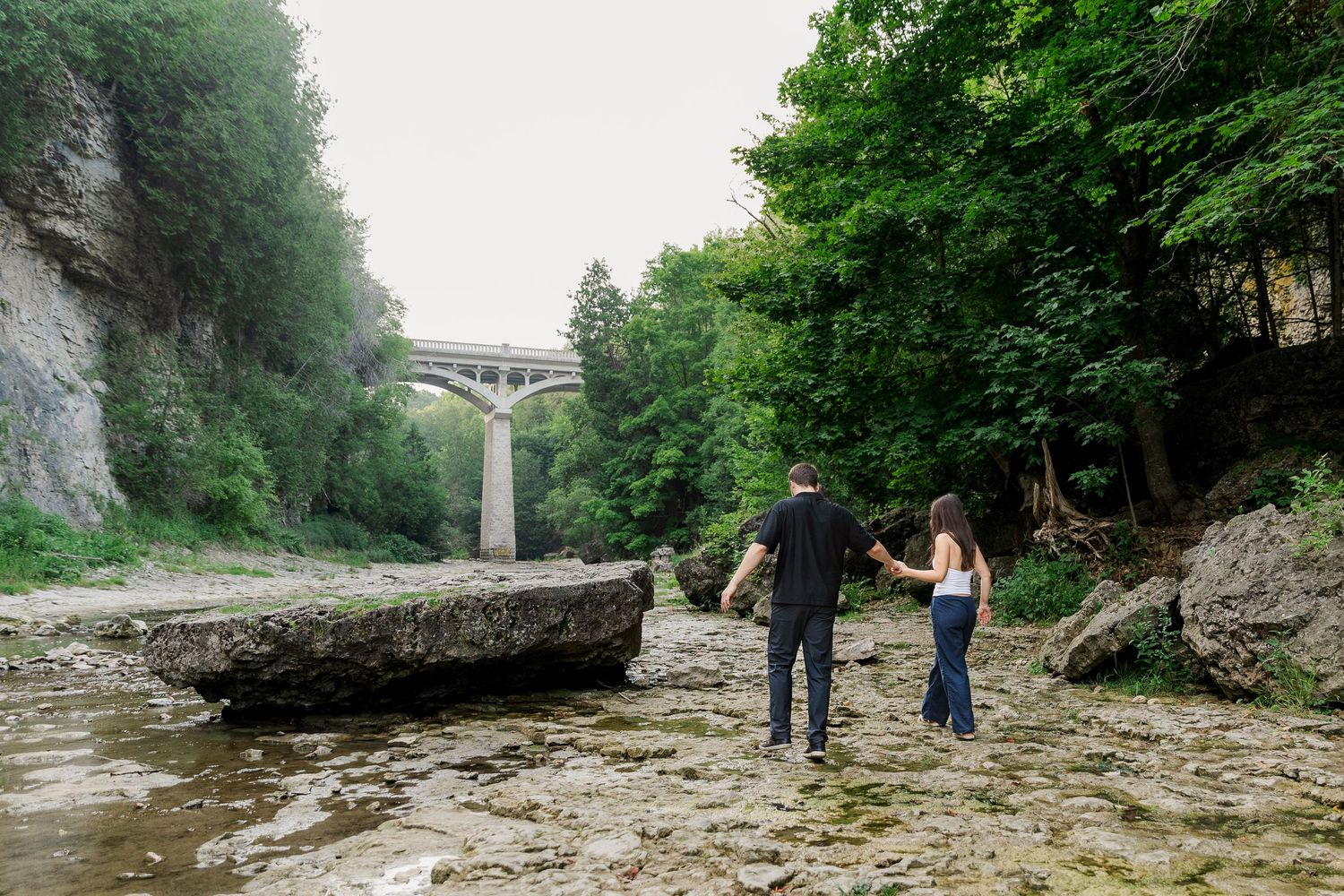 Couple holding hand walking together among the rocks on the river bed of the elora gorge during their engagement session in elora, ontario.