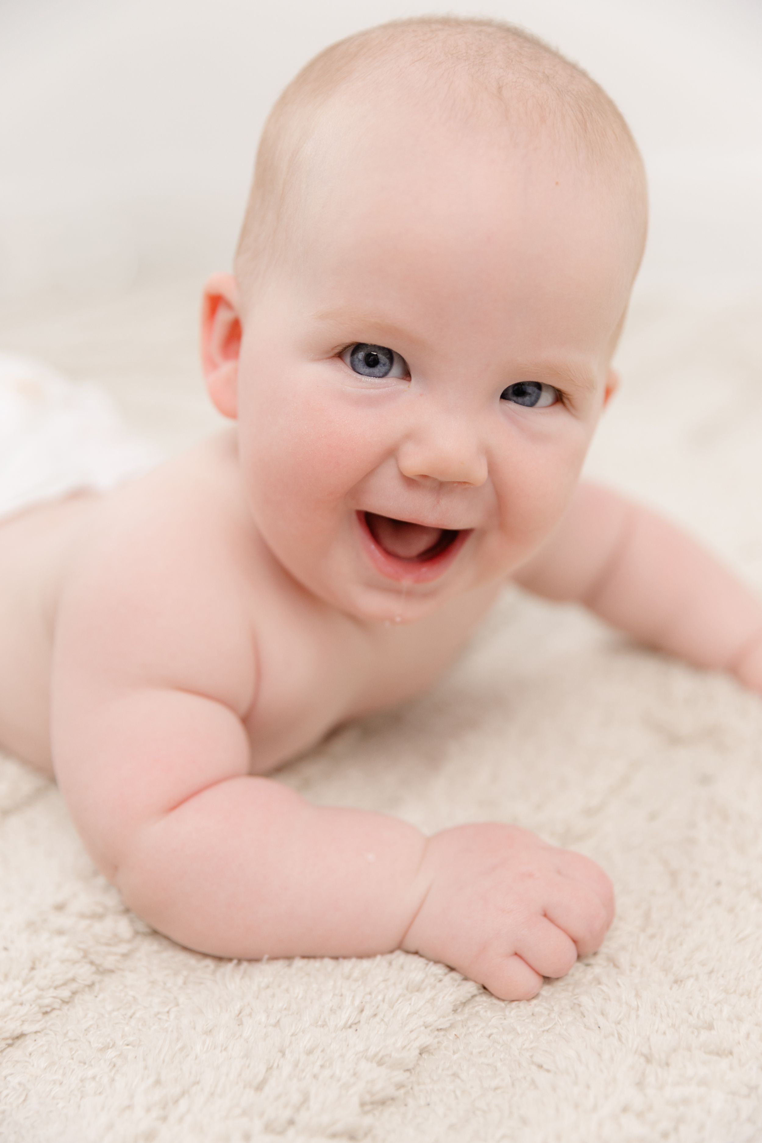 Smiling baby with blue eyes lying on tummy on a soft cream blanket.