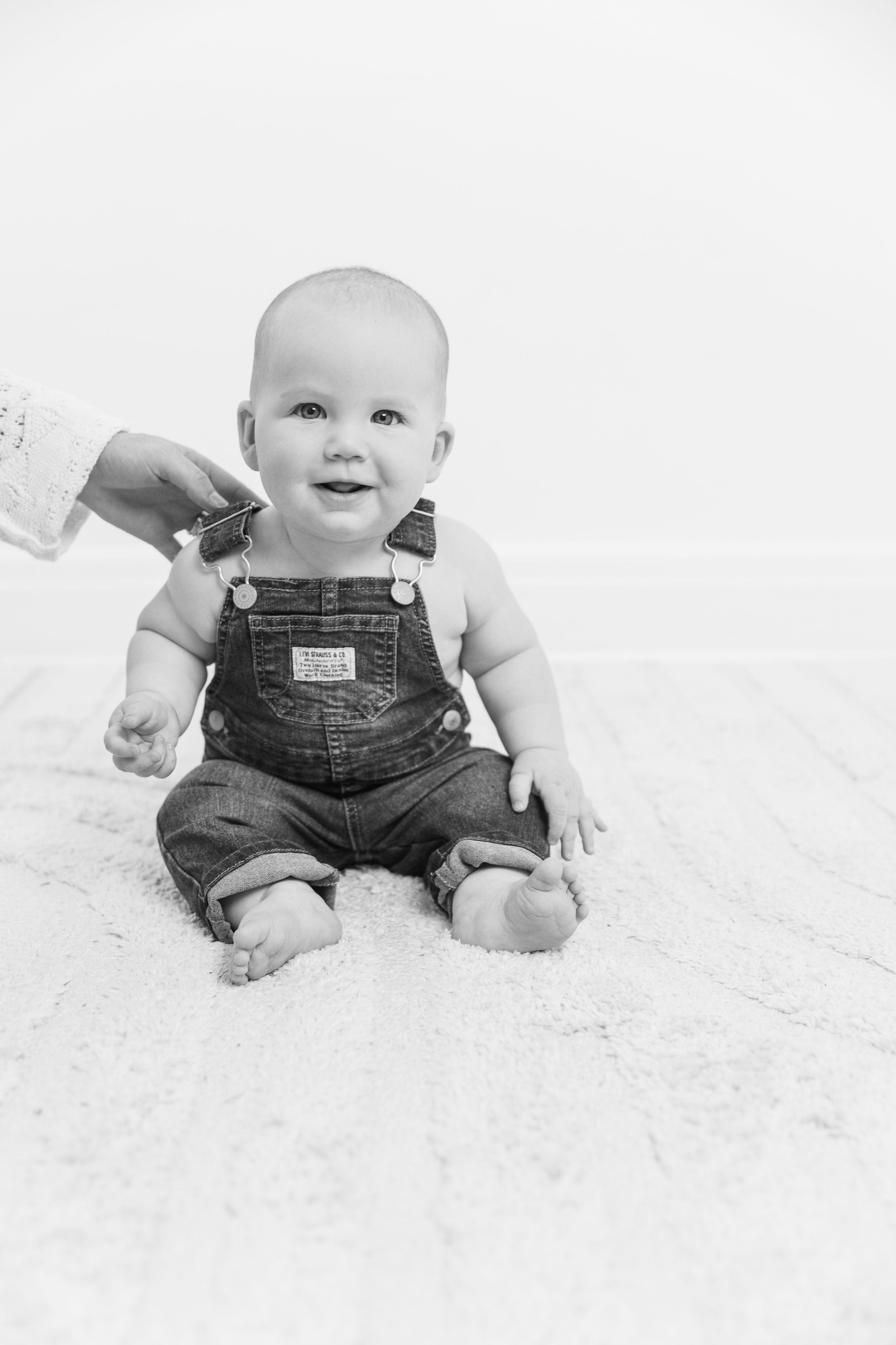 Black and white photo of a smiling baby in denim overalls sitting on carpet with an adult hand visible behind.
