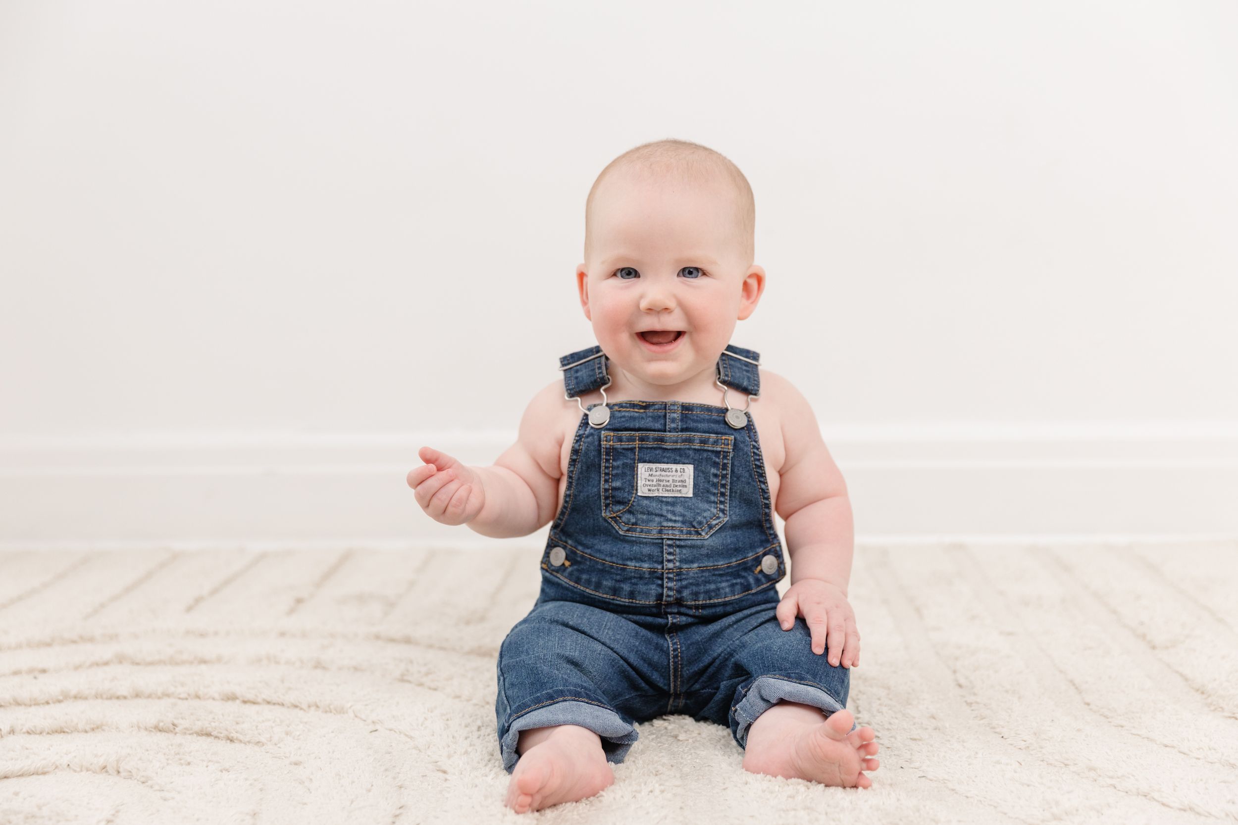 Smiling baby in denim overalls sitting on a white rug against a light wall.