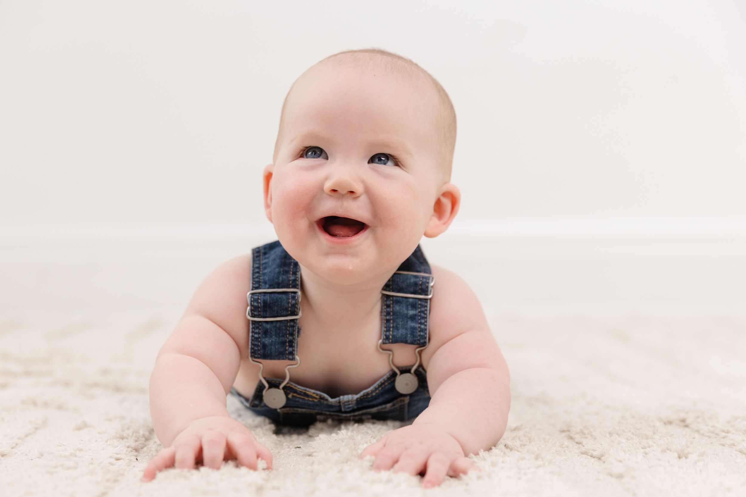 Smiling baby in denim overalls doing tummy time on a soft white carpet, looking up with a joyful expression.
