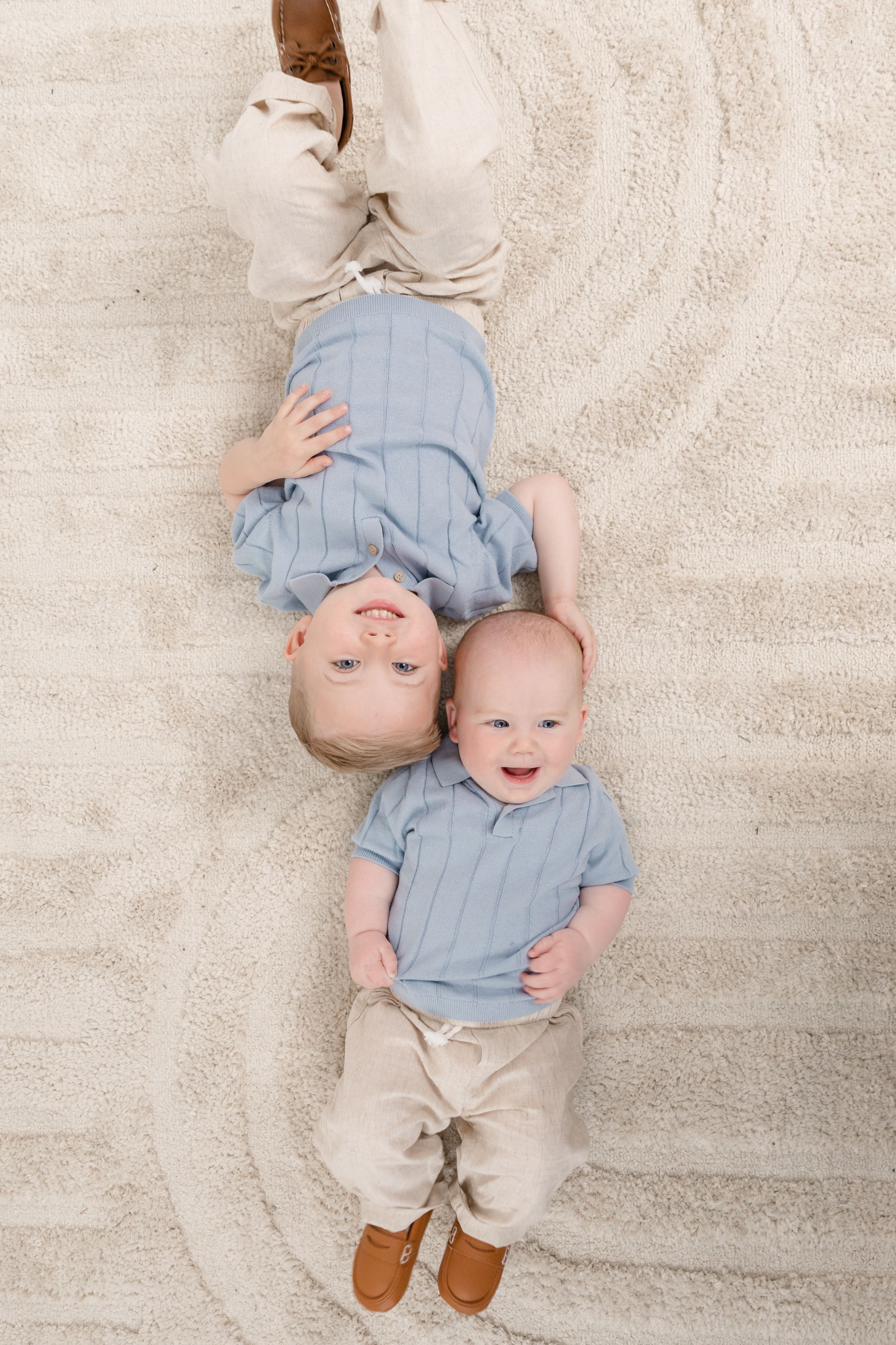 Two young siblings in matching blue shirts lying on a cream carpet, smiling up at the camera.
