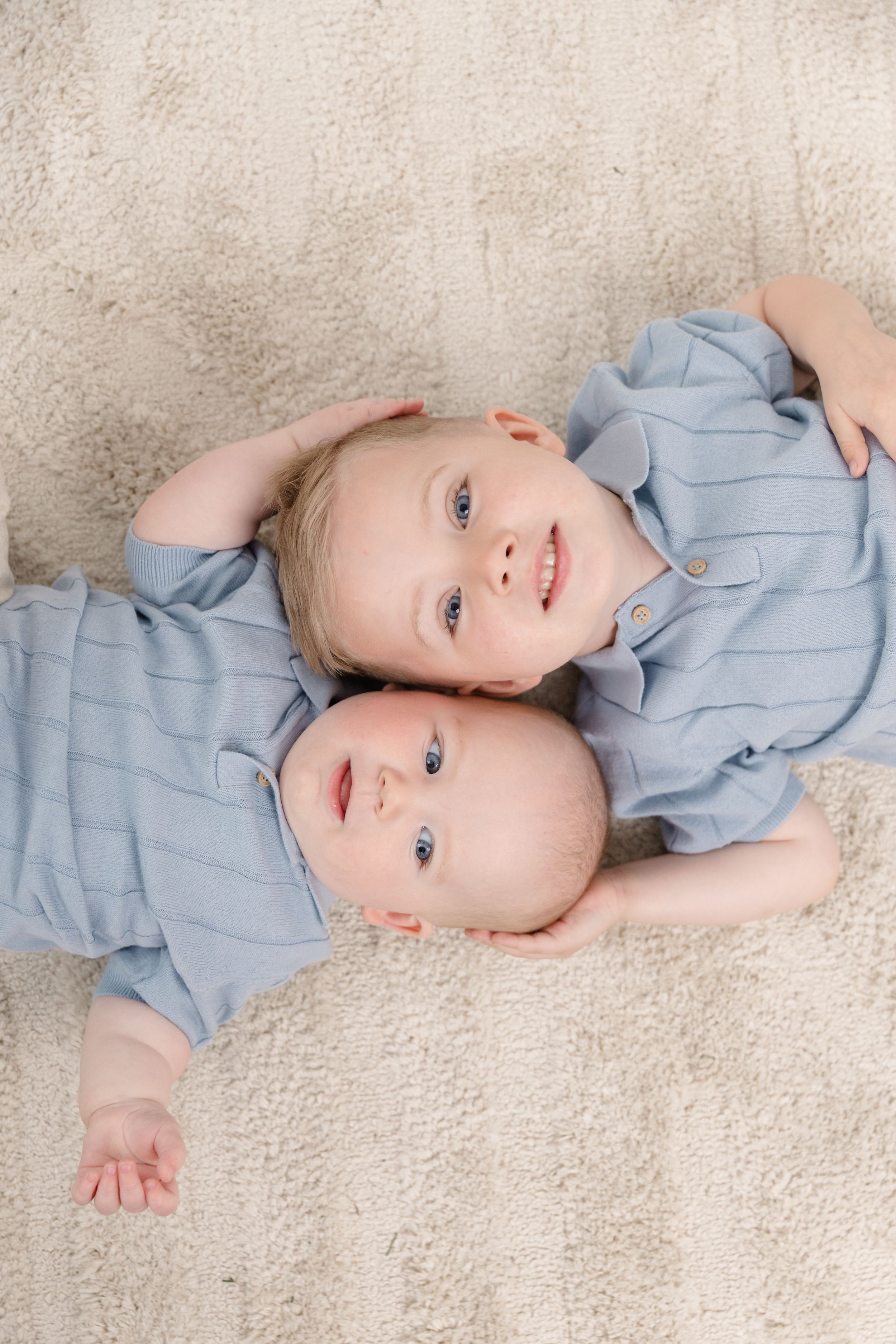 Two young siblings in matching blue outfits lying on a cream carpet, smiling at the camera.